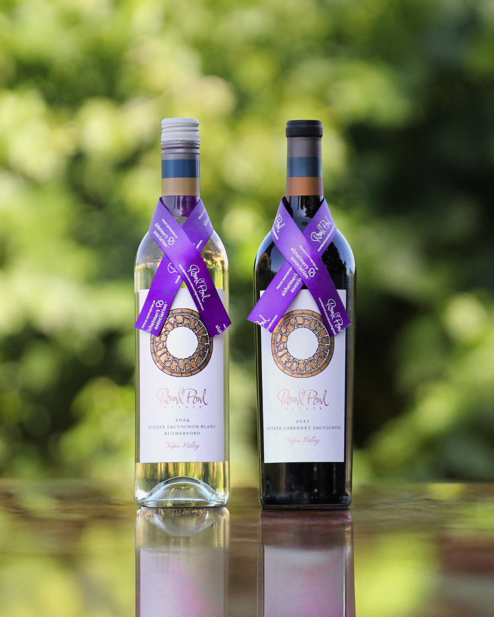 Two wine bottles with purple ribbons, one white and one red, on a reflective surface outdoors.